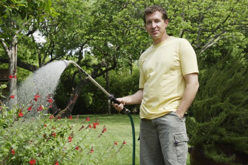 A man stands in a well-maintained garden in Brent Cross, surrounded by lush green trees and shrubs. He is wearing a light yellow T-shirt and grey trousers, holding a garden hose that is watering flowering plants with small red blooms. The garden features a neatly trimmed lawn with dense grass, bordered by flower beds and natural soil areas. In the background, there are mature trees with ample foliage, providing shade and creating a tranquil outdoor environment. The weather appears sunny and clear, ideal for outdoor gardening activities. This scene exemplifies typical garden maintenance and landscaping practices supported by services like hedge trimming and lawn care, contributing to a tidy and healthy outdoor space commonly found in North London suburbs. The overall setting highlights the natural textures of grass, leaves, and flowering plants, capturing an inviting and professional horticultural environment.