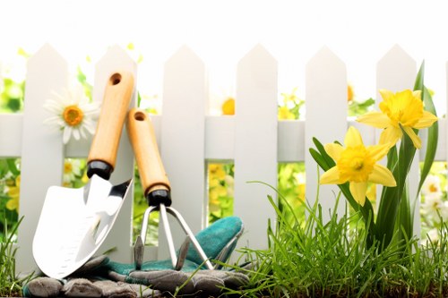 In the foreground of the outdoor garden space, there is a neatly maintained grassy lawn with dense, vibrant green grass. Adjacent to the lawn, a small flower bed contains blooming yellow daffodils with elongated green leaves, adding a splash of bright colour. Behind the flower bed, a white picket fence runs horizontally across the image, providing a charming boundary for the garden. In front of the fence, garden tools such as a trowel and a hand rake with wooden handles are resting on the soil, suggesting recent or ongoing gardening activities. The background hints at a lush, green environment with sunlight filtering through, creating a warm and inviting atmosphere typical of residential outdoor spaces in Brent Cross, London. The garden appears well-kept and suitable for various gardening services offered by Hedge Trimming Brent Cross, including hedge trimming, lawn maintenance, and flower bed care, emphasizing a tidy and aesthetically pleasing outdoor area.
