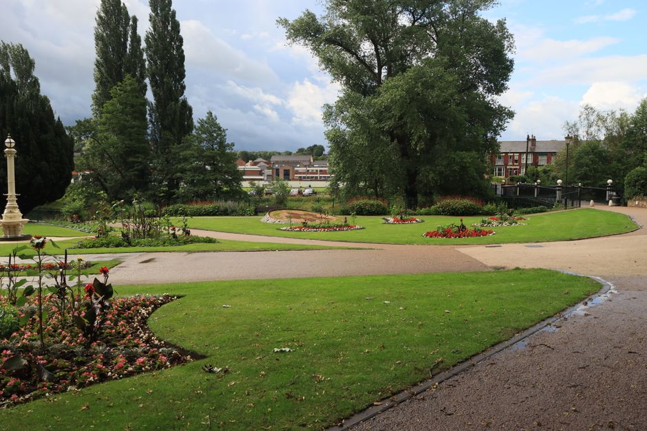 The image depicts a community garden scene at Clitterhouse Recreation Ground, with a gravel pathway running through the centre, bordered on both sides by raised garden beds filled with a variety of yo